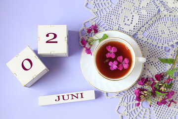 Calendar for June 2: cubes with the numbers 0 and 2, the name of the month of June in English, a cup of tea with purple flowers in it, a gray openwork napkin on a light background, top view