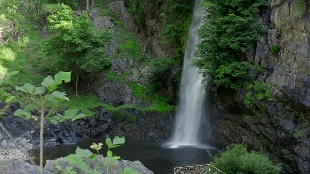 Beautiful landscape with a waterfall in the wild jungle.  Drone flies away from the waterfall, you can see bright green trees, gray stones, sprays of the waterfall and lianas
