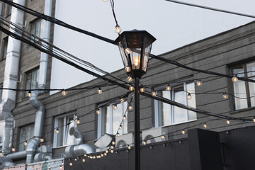 A black lamppost with a garland of lamps on the background of an urban building. City center, modern quarter. Street decoration. A modern city. Windows and metal pipes on the background.