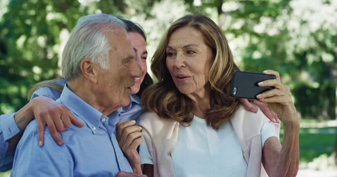 Cinematic shot of happy granddaughter and grandparents having fun to make selfie or technology video call together with smart phone in green park.Concept: connection, communication, generation, family