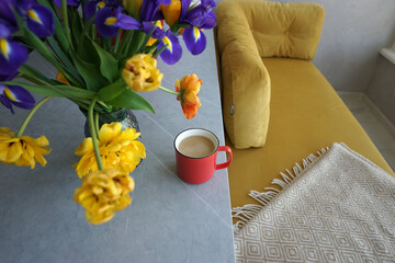 Bouquet of yellow tulips and irises in a vase and a red cup with coffee on a gray table