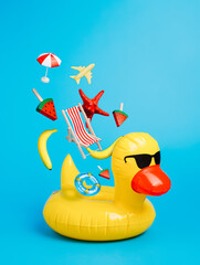 Yellow inflatable floating duck with flying beach chair, starfish, ice cream and and plane on a blue background. Creative trendy summer vacation concept.