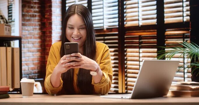Young beautiful Vietnamese student sitting at home and during break in preparation for lectures chatting with friends on social networks holding smartphone.