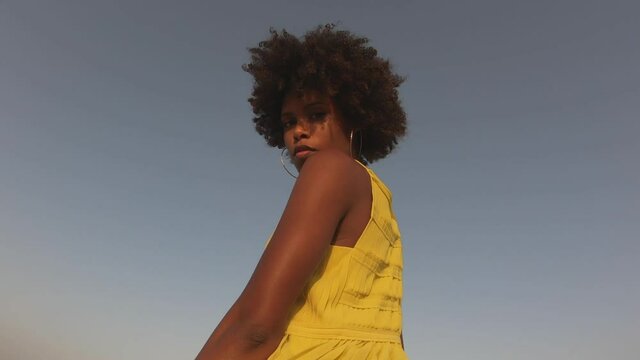 Black woman with afro hair wearing yellow dress against blue sky slowly turning around