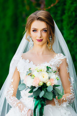 a pensive girl the bride stands before the ceremony with a bouquet of flowers in a park in the middle of greenery.