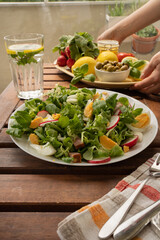 Summer sunny meal with big plate of salad with greens, reddish, eggs, olives. A woman puts a plate with vegetables on the table. 