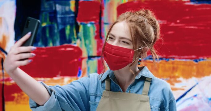 Young cheerful female artist in protective red face mask standing on painting background and posing for selfie on smartphone camera.