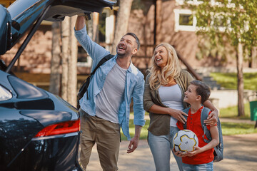 Happy family with little boy packing stuff into the car while standing in front of the house...