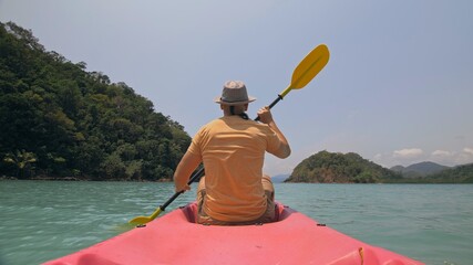 Man with sunglasses and hat rows pink plastic canoe along sea against green hilly islands with wild...
