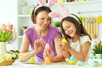  Mother with daughter wearing rabbit ears decorating Easter eggs at home