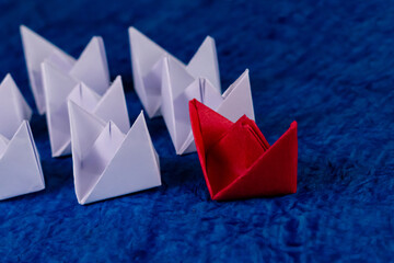 High angle close up shot of one red paper boat with many white boats behind on blue paper as ocean.