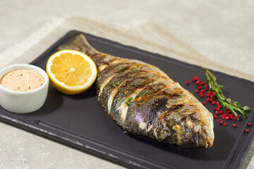 Grilled fish served on a black plate with sauce, lemon and rosemary. horizontal arrangement. Summer menu concept.