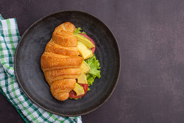 Top view of a croissant with avocados sliced, tomato, and lettuce on a black plate placed on a dark gray background. Space for text. Concept of foods