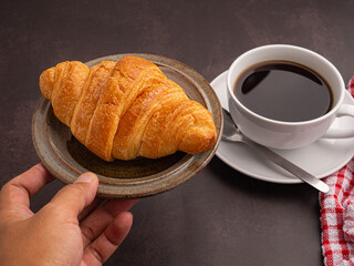 Hand holding of a croissant on a plate with a white coffee cup and a cloth placed on the old kitchen table. Space for text. Vintage background. Concept of dessert, beverage, and relaxation