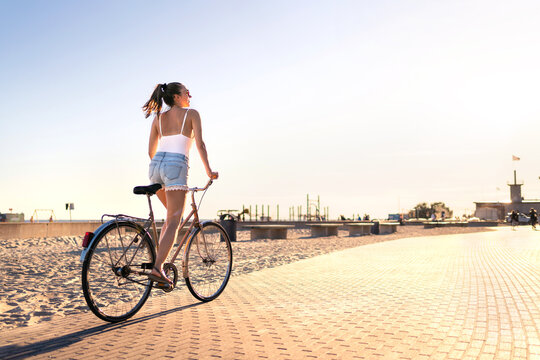 Bicycle fun on beach promenade. Happy woman riding bike on sunny summer boulevard. Seaside waterfront street for cycling. Stylish cyclist lady. Cool carefree feeling at sunset. Summertime freedom.