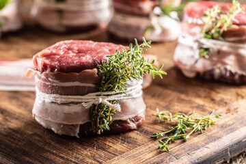 Raw steak wrapped with bacon and fresh thyme on a wooden cutting board.
