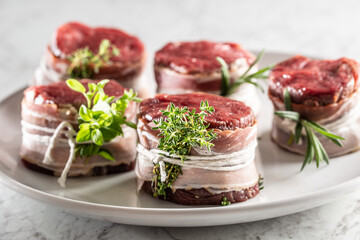 Raw steaks wrapped with bacon and fresh herbs on a plate