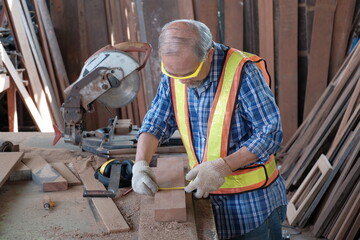 Old Asian man carpenter working in a woodworking factory. They are using a tape measure and a pencil, and other industrial equipment such as hammers, electric saws, and other crafting tools.