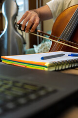 selective focus of a computer a spiral notebook and a young man playing the cello in the living room of his home online class concept