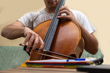 detail of a young man playing the cello at home while taking an online lesson with a notebook and computer while in confinement.