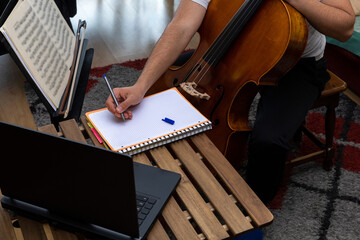 young musician taking notes in a notebook on a wooden table in an online class with cello laptop...