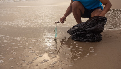 Blue mask Someone is picking up the stick. About to put on a black bag by the sea, water washed up concept seaside garbage, masked garbage, covid-19