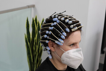 young woman, girl is getting perm,  hair curlers on head at hairdressers salon, wearing mask...