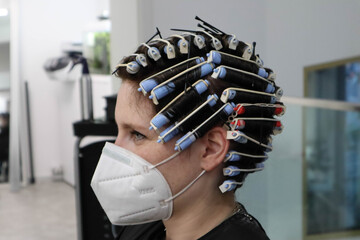 young woman, girl is getting perm,  hair curlers on head at hairdressers salon, wearing mask...