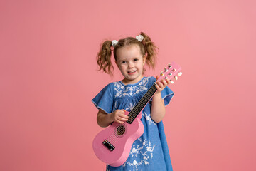 cheerful little Caucasian girl of 5 years old in denim dress plays pink ukulele. child plays a musical instrument and sings on a pink background. concept of summer and the beginning of the holidays