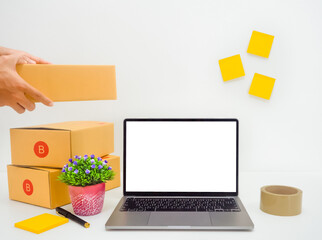 A white notebook screen placed in the center, someone holding a parcel in the upper corner. Next to it is a brown box, masking tape, a small tree, pen with white background. Online sales concept.