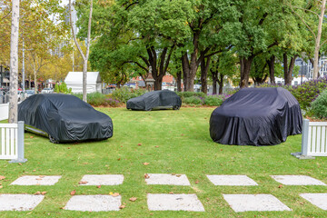 Three mystery cars are draped under black covers in a park setting in Melbourne Australia
