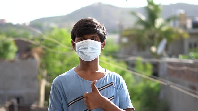 Portrait of an Indian teen giving thumbs up to the camera while wearing face mask and standing outdoors