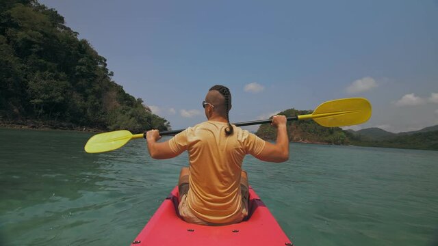 Strong man with thin plait hairstyle rows pink plastic kayak putting up paddle on sea against hills with wild jungles and blue sky. Young guy is sailing on kayak in ocean. Traveling to tropics.
