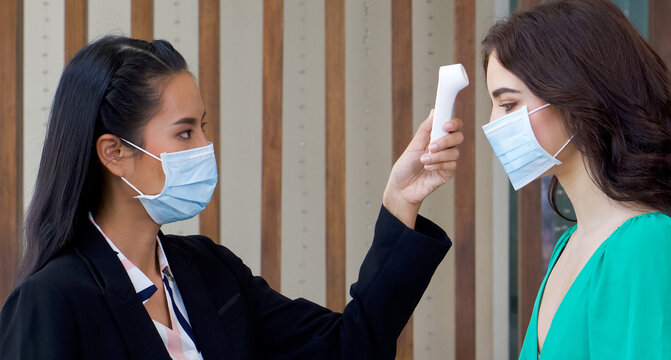 Asian woman in black suit measuring the temperature of caucasian woman in green dress with a fever monitor diagnostic tool. Both wear face mask to protect from the coronavirus outbreak.