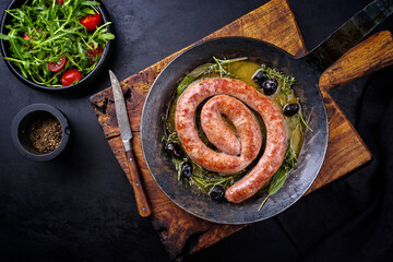 Traditional fried Italian salsiccia fresco meat sausage served with rocket salad and olives as top view in a cast iron pan