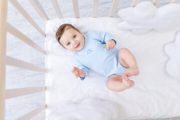 happy smiling baby boy in crib in blue bodysuit, cute joyful little baby in bedroom