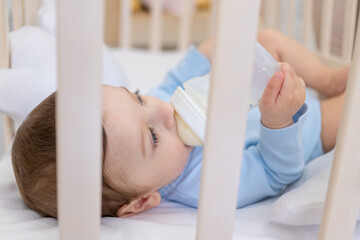 baby boy eating from milk bottle in crib in blue bodysuit, cute little baby in bedroom, baby food concept