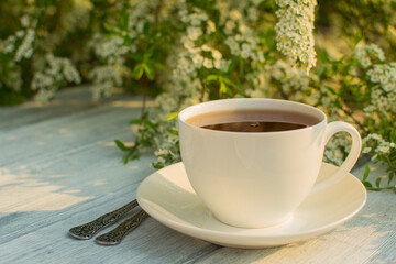 White porcelain cup with tea on a wooden table against a background of white flowers.