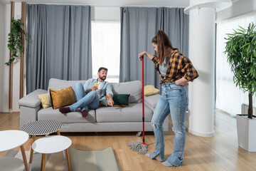 Young couple having fun at their home where man making fun with his girlfriend who cleaning and...