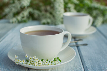 Two white porcelain cups with tea on a wooden table against a background of white flowers.