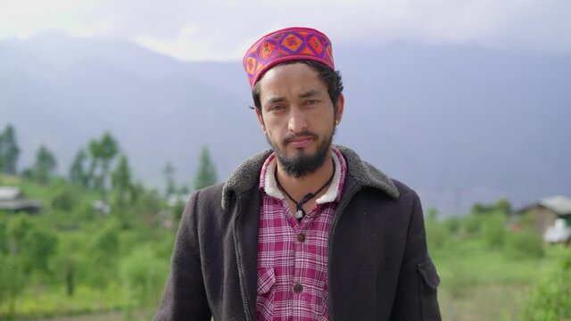 A young Indian male Himachali man (Pahari male) wearing a traditional or ethnic cap with mighty Himalayan Mountain range in the background looking at the camera in a broad daylight 