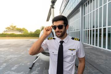 Young male pilot standing next to his helicopter ready to take off. He is in his uniform with sunglass. The helicopter is parked outside a hanger.