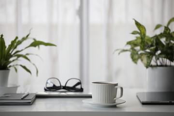 Ceramic white coffee cup and black glasses with laptop and plant pot on white desk