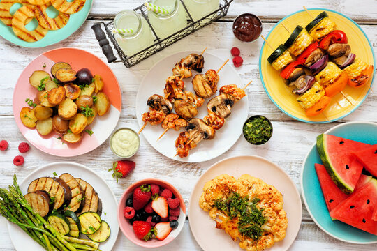 Vegan summer bbq or picnic table scene. Overhead view on a white wood background. Fruit, grilled vegetables, skewers, cauliflower steak and lemonade. Meat substitute concept.