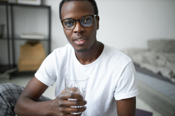 Close up image of attractive young dark skinned guy with stubble holding glass of water, refreshing himself after awakening or morning workout, feeling thirsty. Daily routine, habits and hydration