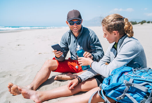 Teenager Boy passing drinking water bottle to Smiling Father in sunglasses with smartphone sitting with backpacks on the sandy seaside beach. Active happy family people vacation concept image