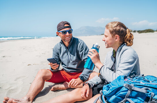 Smiling Father in sunglasses with smartphone sitting with teenager son with backpacks on the sandy seaside beach. Boy drinking water from bottle. Active happy family people vacation time concept image