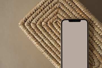 Flatlay of blank screen mobile phone on rattan casket and neutral beige background. Home office...