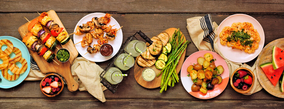 Healthy plant based summer bbq table scene. Overhead view on a dark wood banner background. Grilled fruit and vegetables, skewers, cauliflower steak and vegetarian sides. Meat substitute concept.