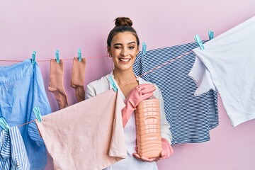 Beautiful brunette young woman doing laundry holding detergent bottle winking looking at the camera...
